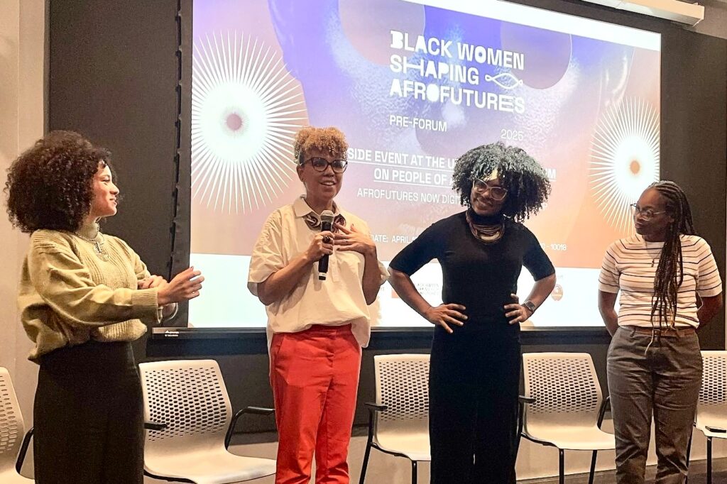 Four Black women standing. One is holding a microphone and the other three are looking at her. In the background is a screen that says; " Black women shaping afrofutures Pre-Forum. 