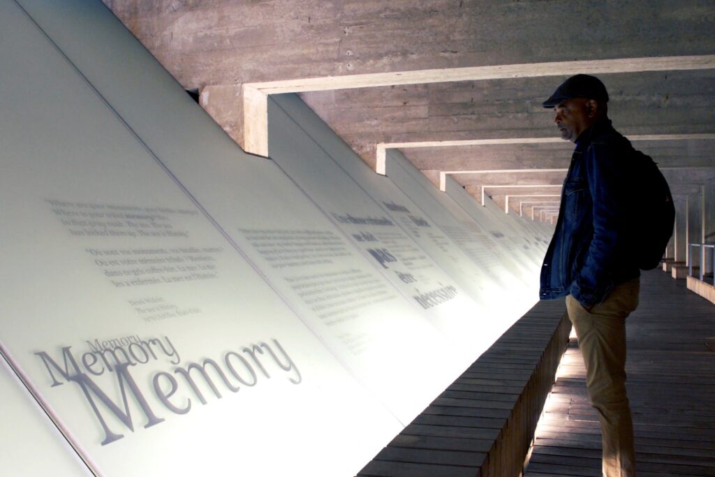 A man standing in front of a lighted board at a museum and on that lighted board it says "Memory, Memory, Memory" as well as a bunch of other wording. 