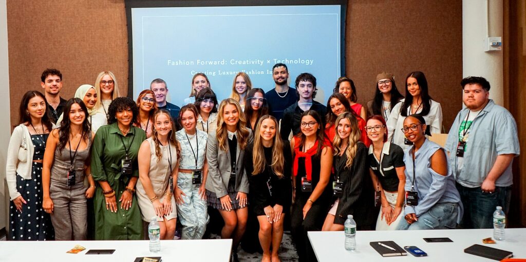 A large group of students and panelists pose together indoors in front of a presentation screen that reads “Fashion Forward: Creativity × Technology.” They smile at the camera, wearing event badges.