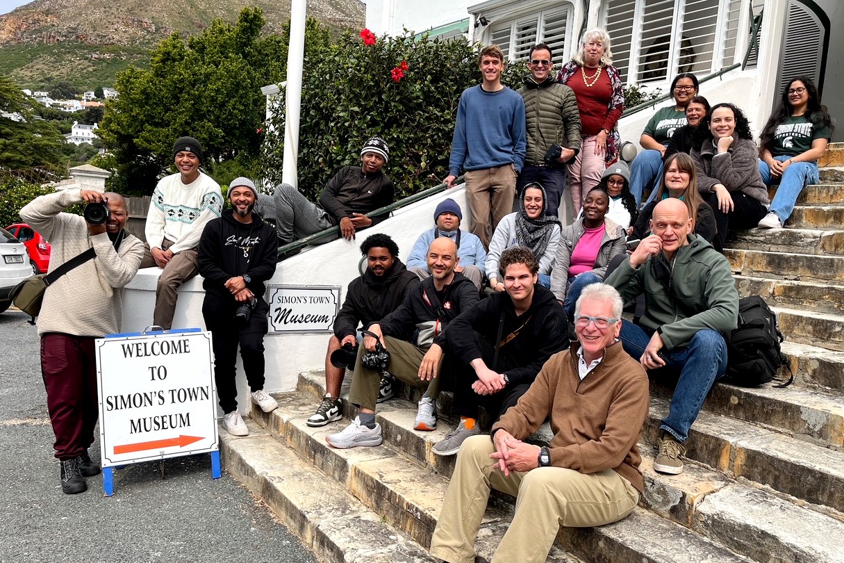A large group, including Peter Glendinning, sit and stand on the stone steps outside Simon’s Town Museum in South Africa. A sign reading “Welcome to Simon’s Town Museum” is visible at the bottom of the steps.