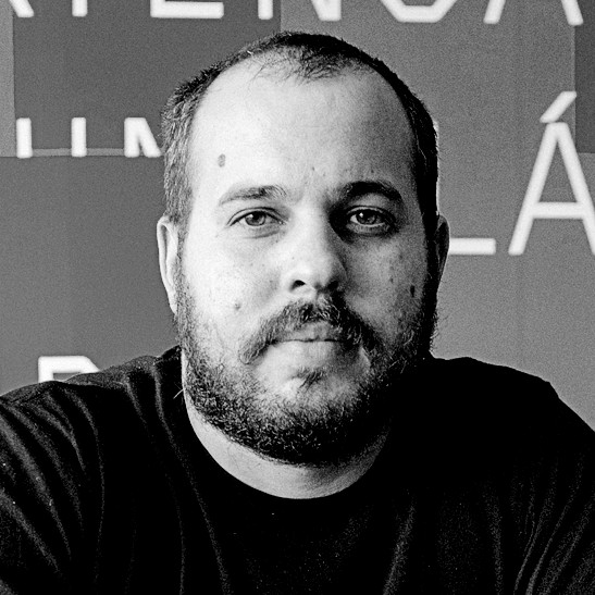 Black-and-white image of a man with a short beard and closely cropped hair, wearing a dark T-shirt. He looks directly at the camera with a neutral expression; text is partially visible on the wall behind him.