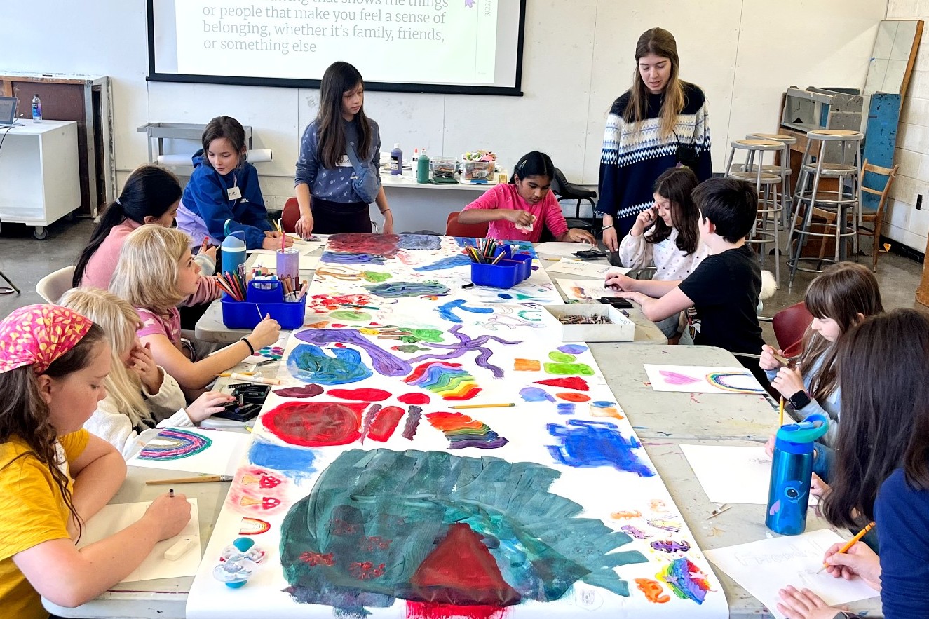 Children sit on both sides of a long table, contributing to a shared painting. The paper canvas is filled with colorful shapes, swirls, and rainbows. An instructor oversees the activity.