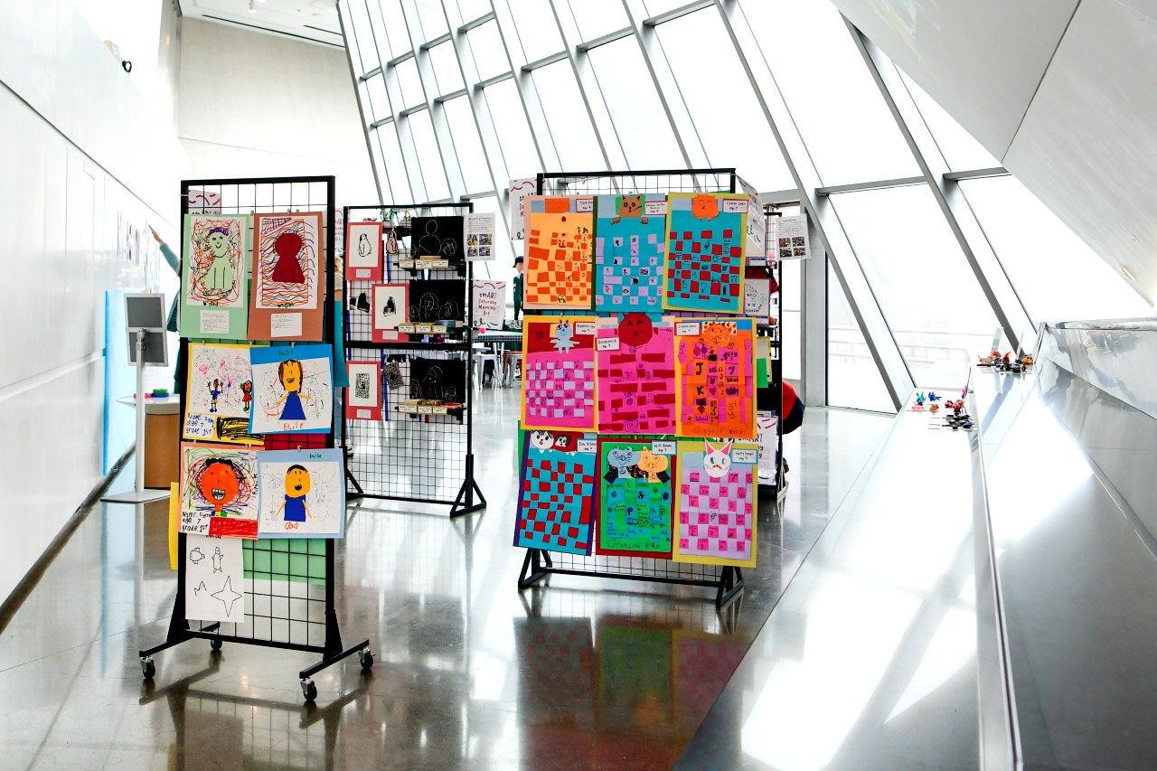 An art exhibit display in a modern gallery space, with several rolling panels filled with children’s artwork. The panels showcase crayon portraits, woven paper collages, and bright, abstract designs. Natural light streams in through the tall glass windows lining the wall.
