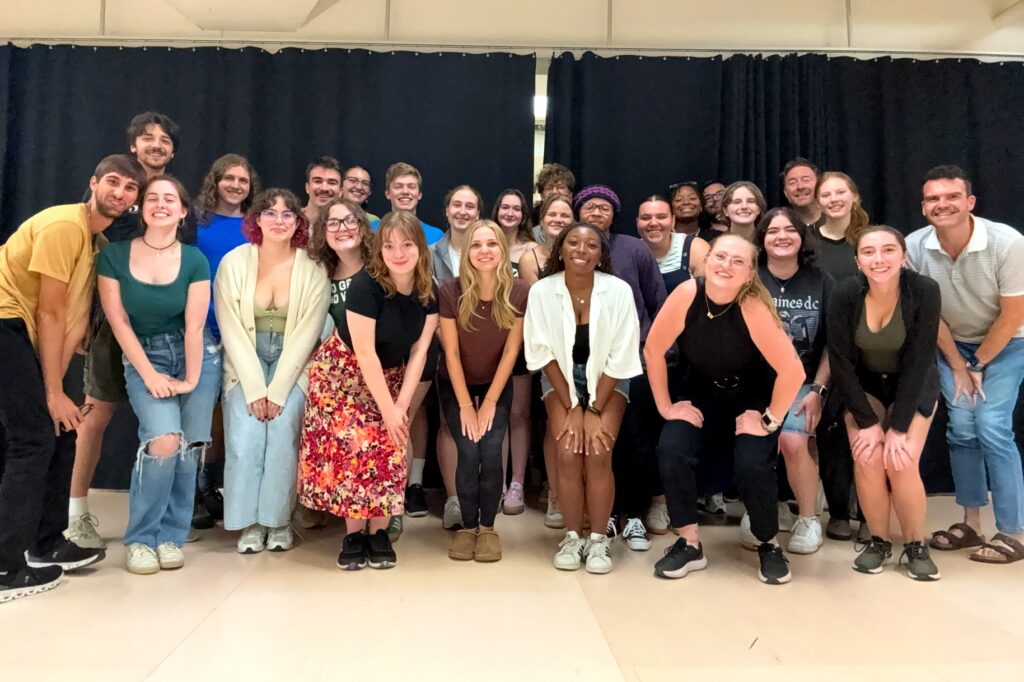 A large group of approximately 30 young adults pose together in a rehearsal space. They are smiling and standing in staggered rows in front of a black curtain backdrop. 