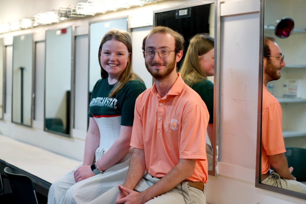 Two students sit side by side on a counter in a dressing room lined with mirrors. The student on the left wears a dark green Michigan State t-shirt over a white corset-style costume piece. The student on the right wears an orange collared shirt with glasses and beige pants. Both smile. 