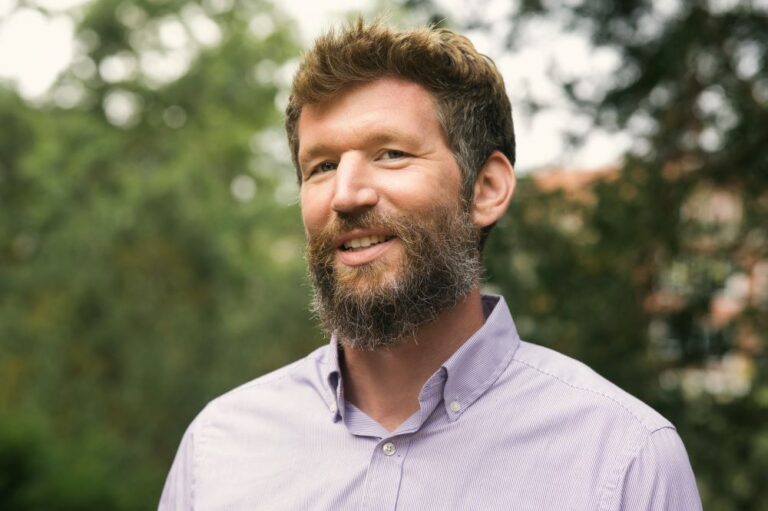 Portrait of MSU Assistant Professor Tim Conrad smiling. He’s wearing a button down short and in the background are tree branches.