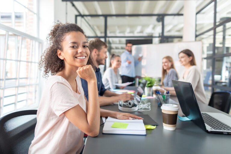A young woman smiles at the camera while seated at a conference table with notebooks and laptops, as colleagues collaborate in the background