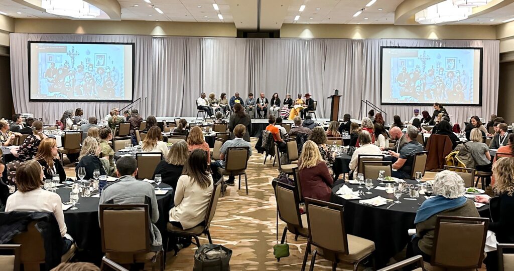 Audience members sit at round tables in a hotel ballroom watching a panel discussion on stage during the Conference on Community Writing. Two large screens flank the stage, displaying a projected image.