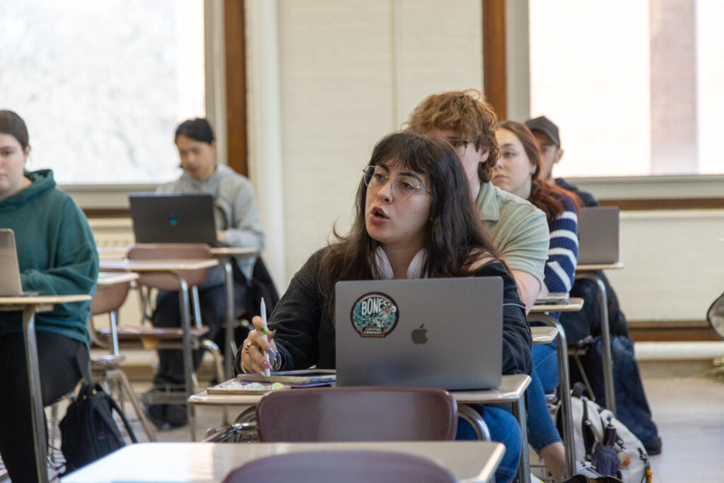 A student speaks during class while holding a pen and sitting behind a laptop with stickers. Other students sit at desks with laptops in the background.