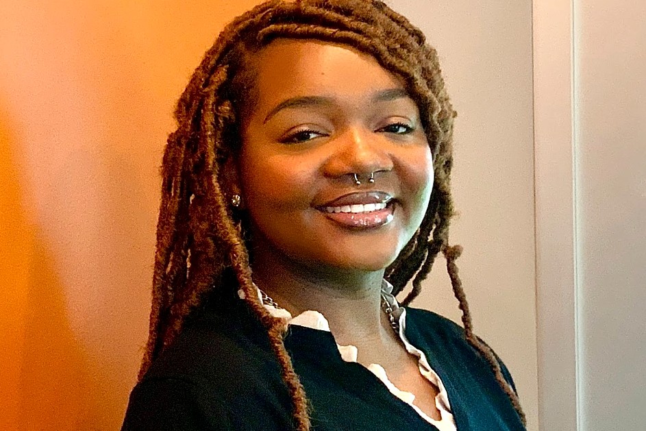 Head and shoulder photo of a Black woman who is smiling and wearing a black V-kneck shirt with white accent around the v-kneck. She has a nose ring and dreadlocks. There is and orange background and in the top left corner is says "Arts4U."