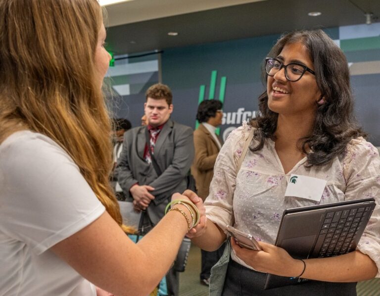 A student smiles while shaking hands with another person during a networking event, holding a tablet and phone. Other attendees in professional attire are visible in the background.