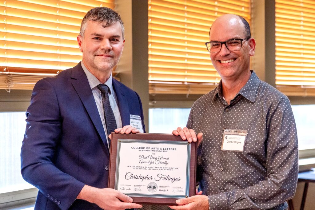Two men stand side by side indoors in front of windows, holding a plaque that reads Paul Varg Alumni Award for a Faculty – Christopher Frilingos, College of Arts & Letters, Michigan State University.”Paul Varg Alumni Award for a Faculty – Christopher Frilingos, College of Arts & Letters, Michigan State University.” The man on the left wears a navy suit and tie, while the man on the right, wears glasses and a patterned button-down shirt. 
