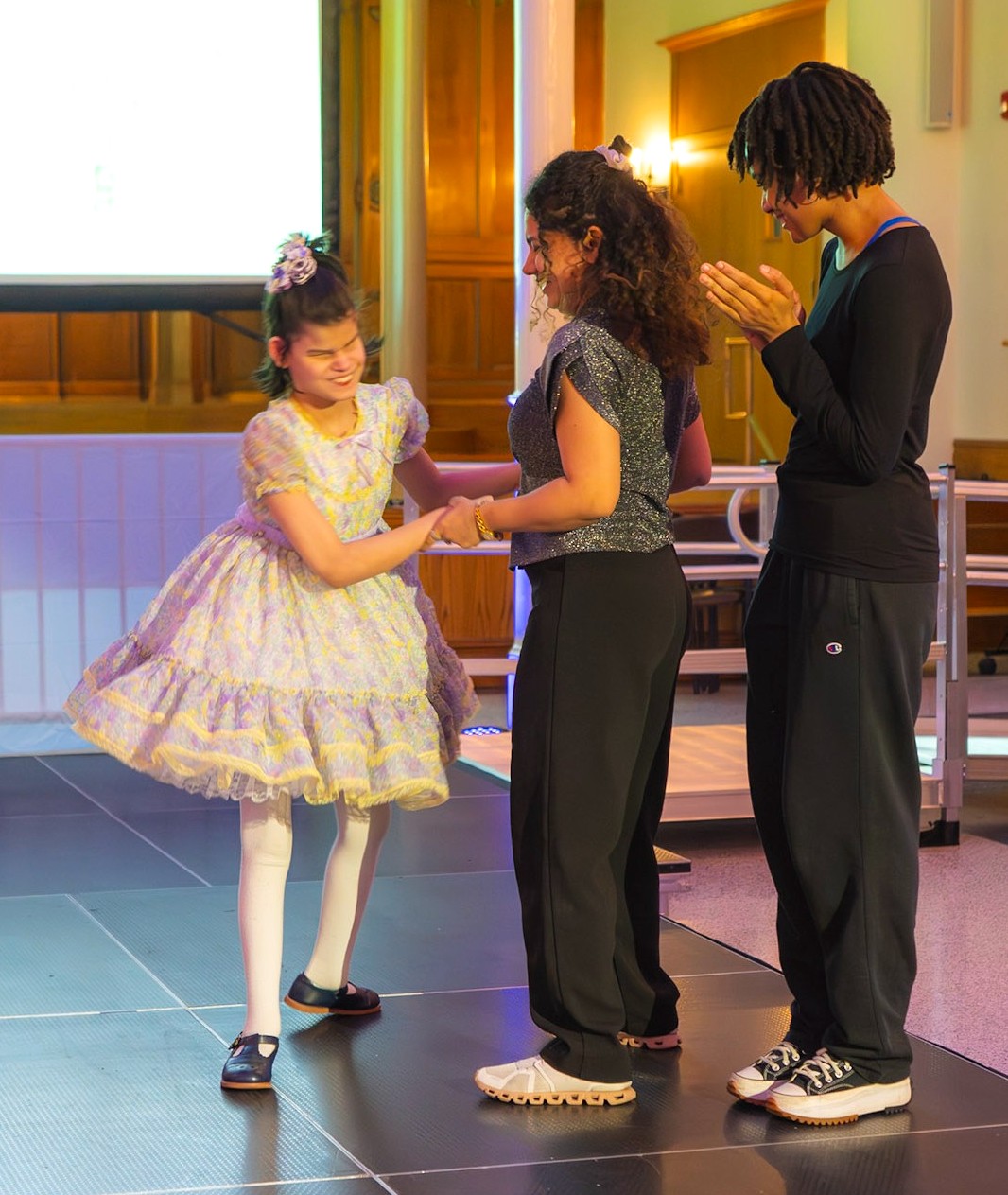 A young girl in a frilly lavender dress stands with another person in a glittery top and black pants, while a third person stands nearby clapping. They are on a runway.