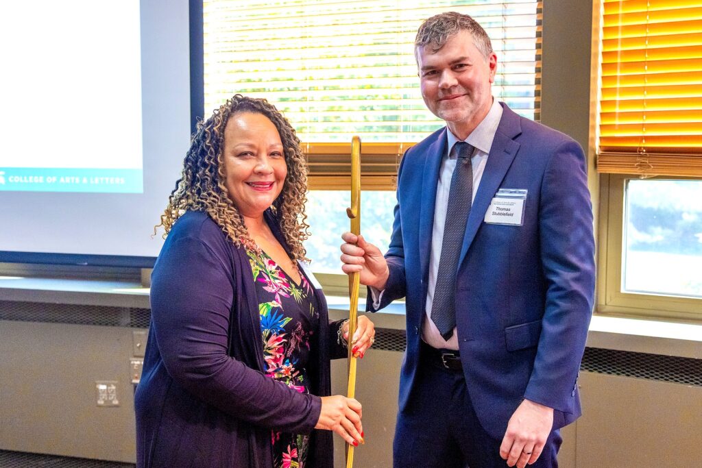 A woman in a colorful floral dress with a dark cardigan smiles while holding one end of a golden staff. A man in a navy suit and tie holds the other end. They are standing in front of a window with natural light coming through the blinds.