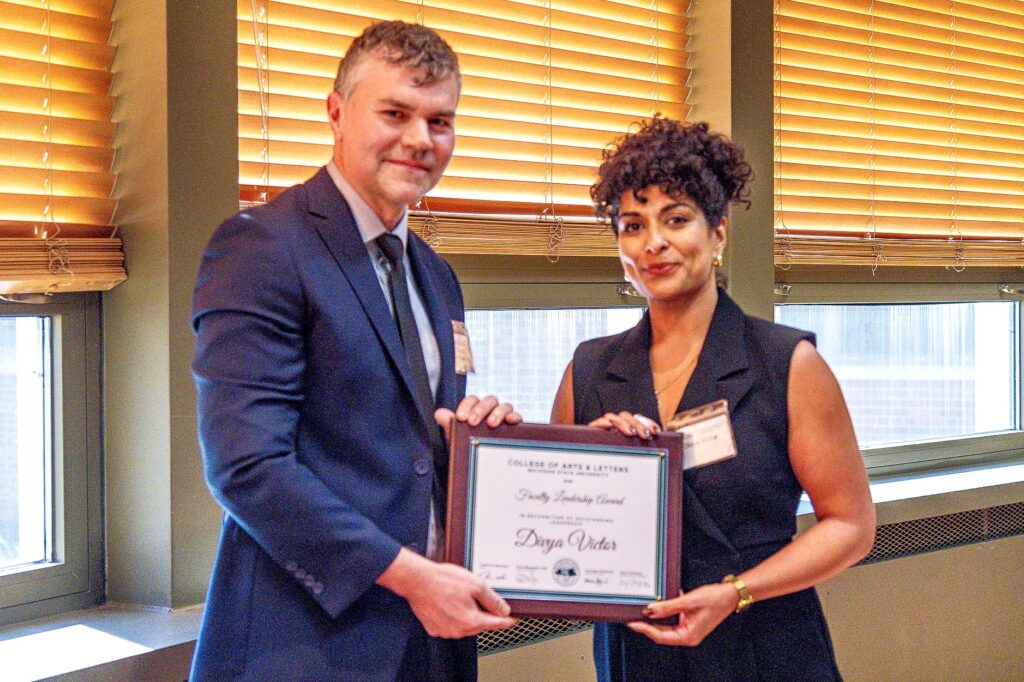 A man in a navy suit and tie stands next to a woman in a black sleeveless blouse. Both hold a framed award and face the camera with small smiles. The plaque reads, “Faculty Leadership Award – Divya Victor, College of Arts & Letters, Michigan State University.”