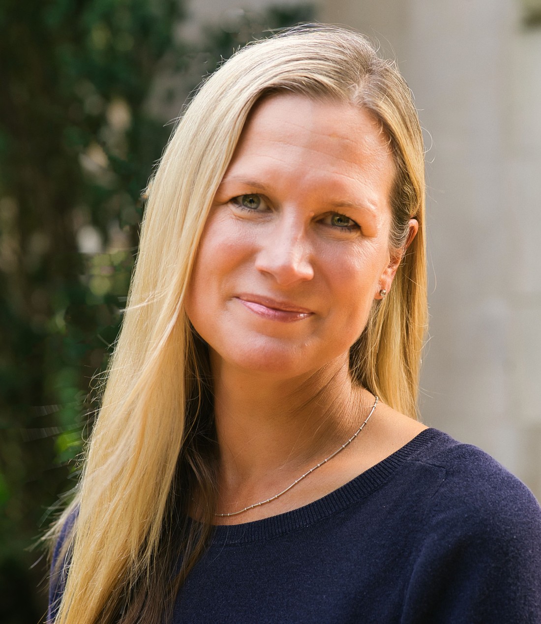 A portrait of a person with long blond hair wearing a dark sweater, standing outdoors with greenery softly blurred in the background.