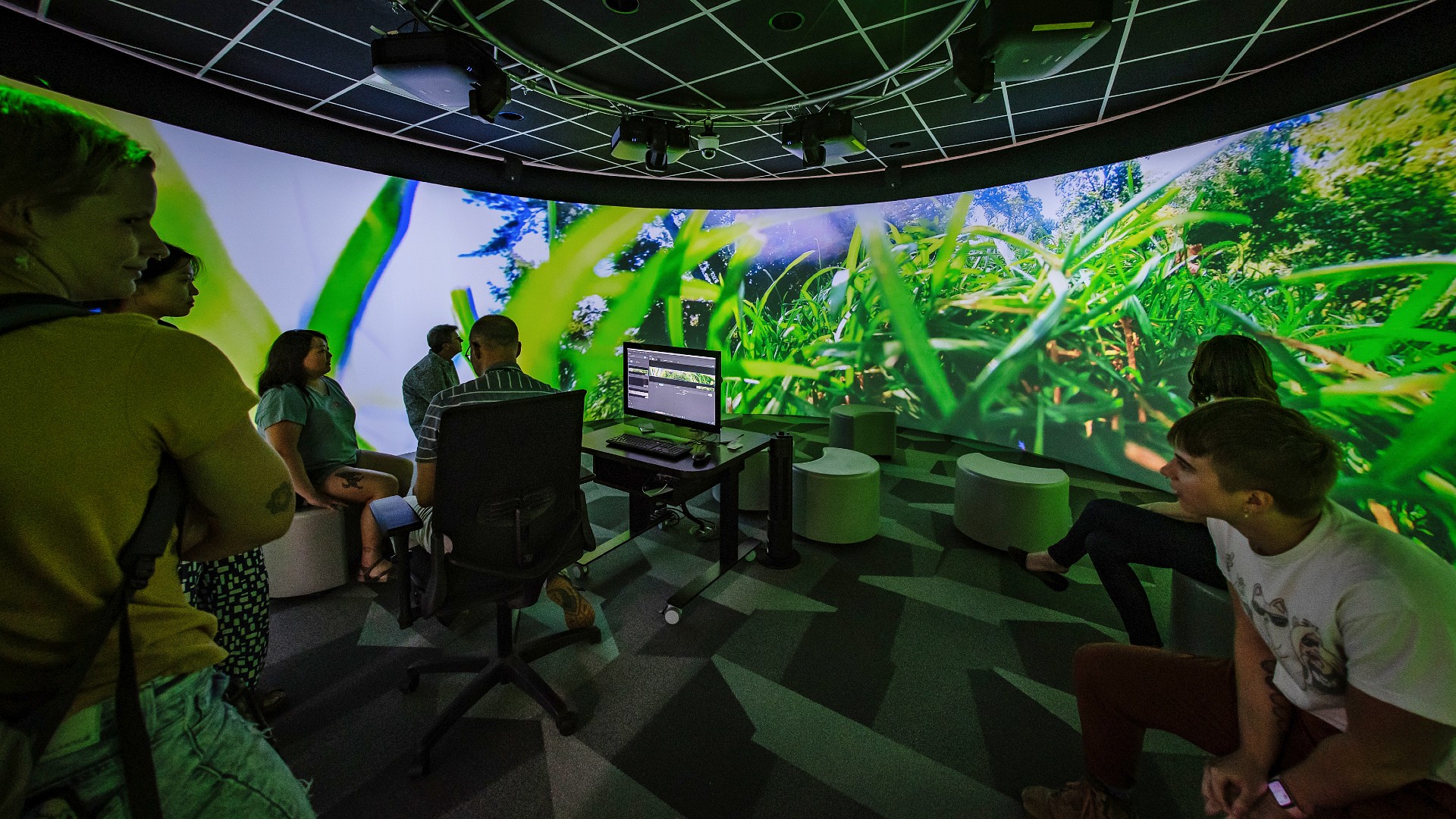 Audience members sit in the MSU Libraries Digital Scholarship Lab’s 360 Room as panoramic projections of grass and trees fill the curved walls during Hailey Becker’s “Where the Light Gets In” virtual tour.