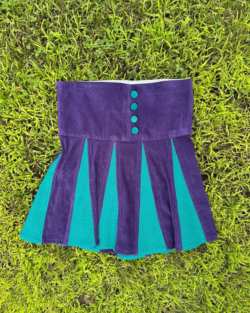 A short skirt made of alternating panels of dark purple and teal fabric with teal buttons on the waistband is laid flat on grass.