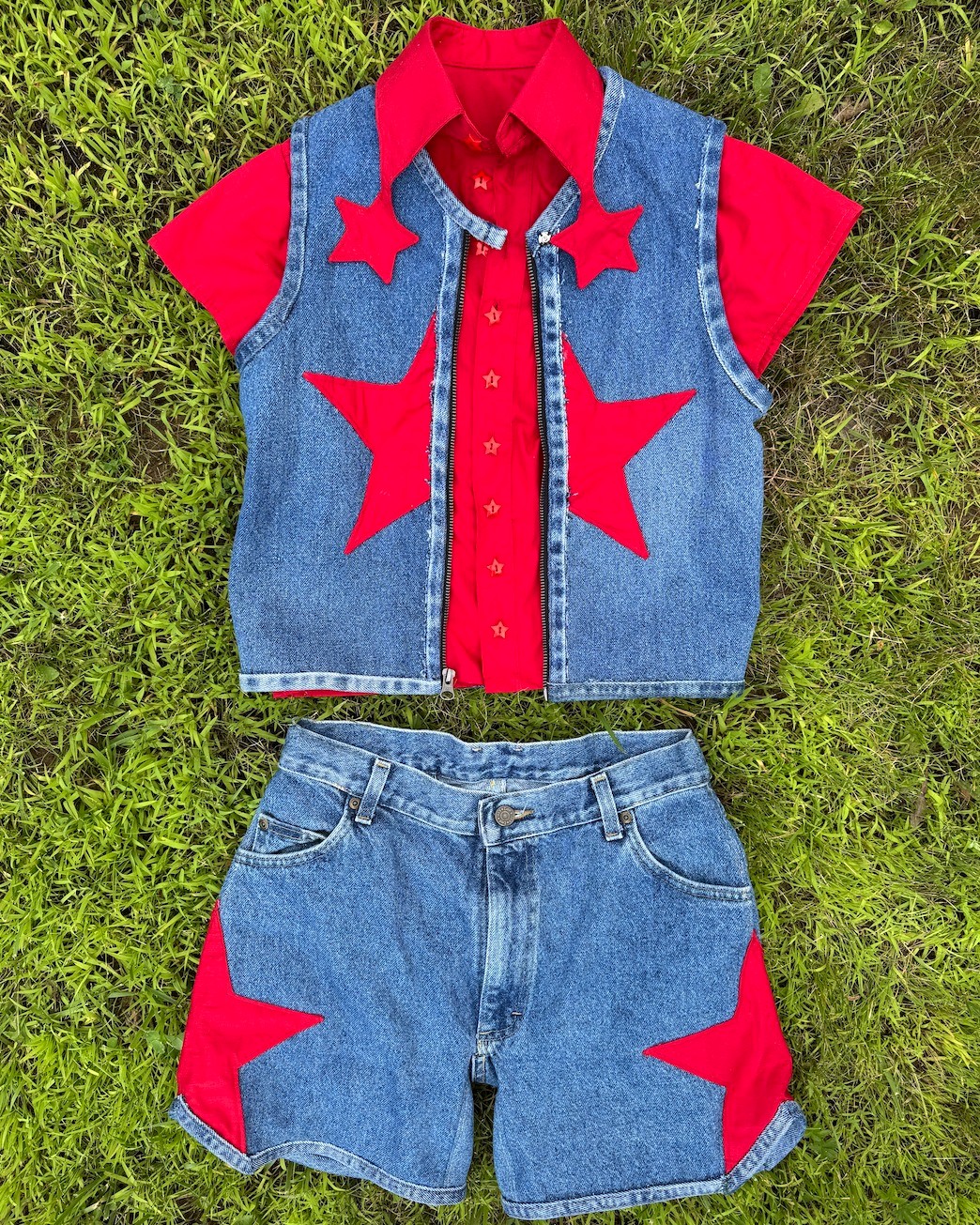 A denim vest and shorts set with red star-shaped appliqués and a red shirt underneath is laid flat on grass.
