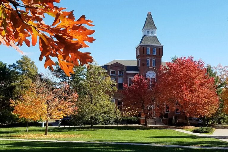 A fall photo of Linton Hall with red fall leaves, green grass and the red brick Linton Hall building with a white wood steeple.