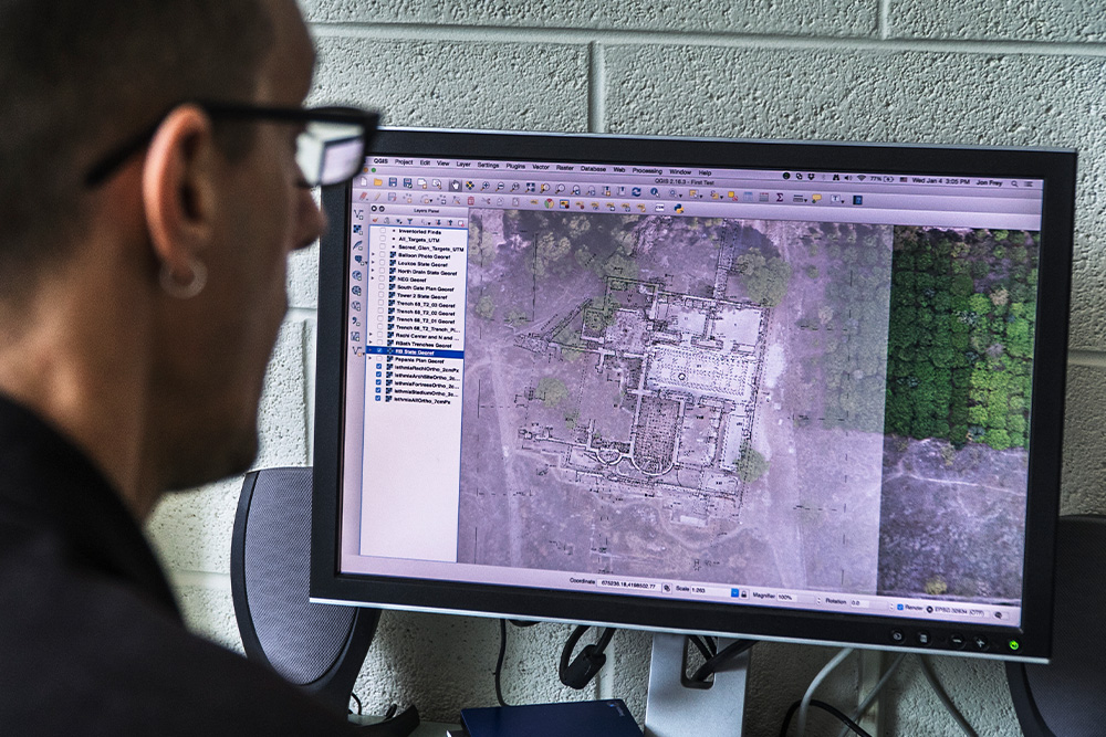 A person in glasses looks at a computer screen displaying satellite imagery and map data of a site with trees on the right. The setting is analytical and focused.