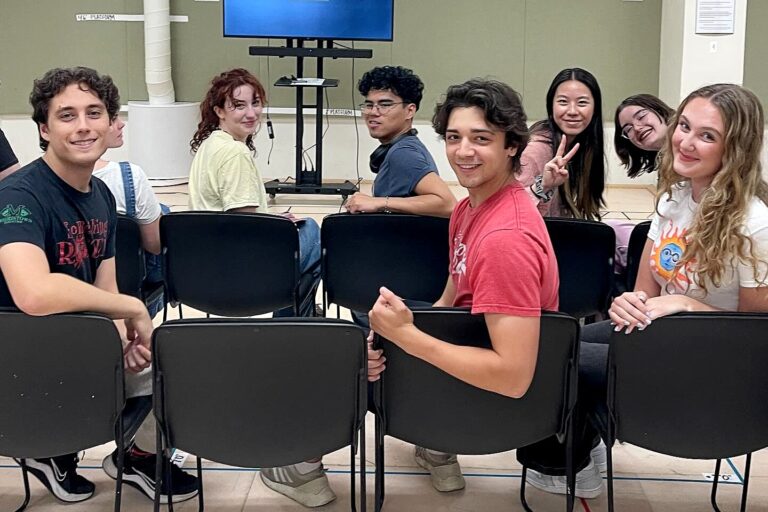 A group of students sit in rows of chairs, turned toward the camera and smiling during a rehearsal presentation.