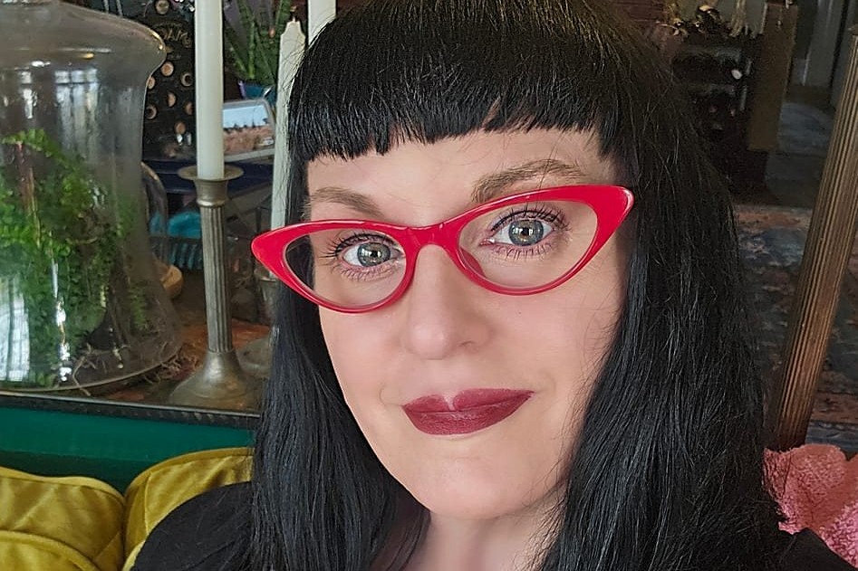 A person with long dark hair and blunt bangs wearing bright red cat-eye glasses and red lipstick, seated indoors with plants and candles visible in the background.