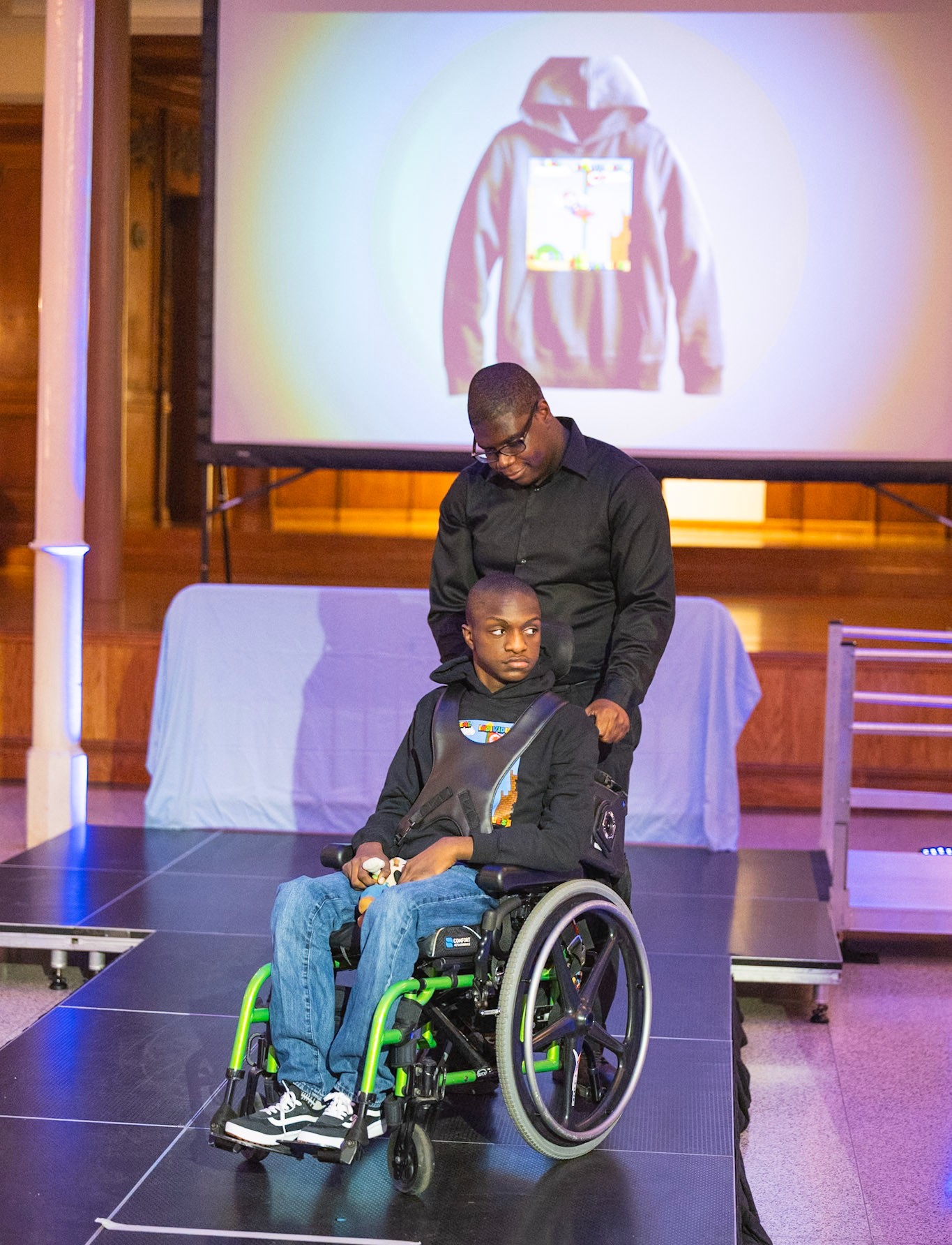 An adult stands behind a child in a wheelchair on a runway, both wearing black clothing. The child wears jeans and a black hoodie featuring a colorful design on the chest. Behind them, a projected image of the hoodie is displayed.