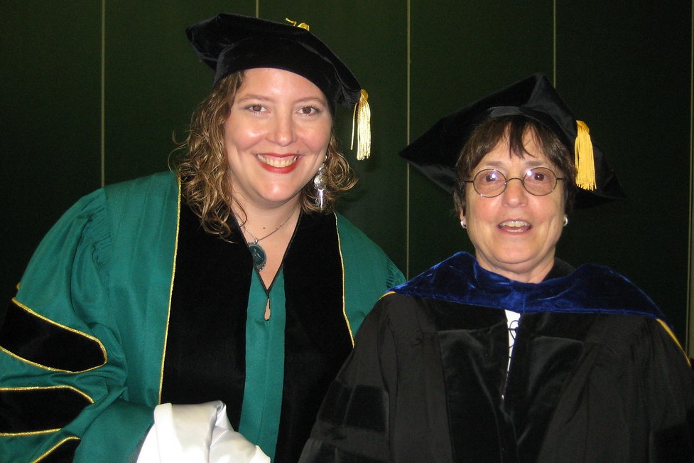 Two people dressed in doctoral regalia stand indoors against a dark green background. The individual on the left wears a green gown with black velvet panels, while the person on the right wears a black gown with a blue velvet hood. Both wear academic caps with gold tassels and are smiling.