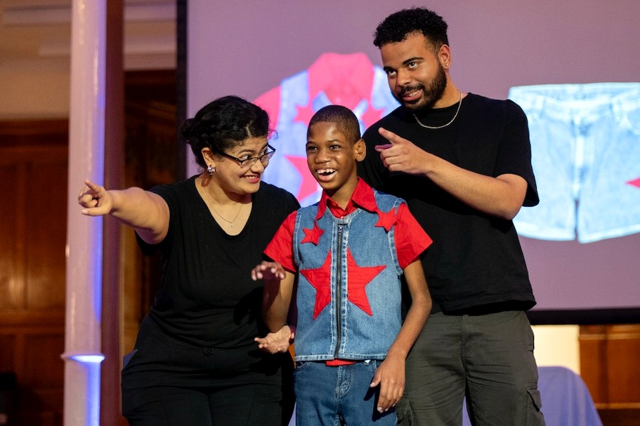 A smiling child wearing a red shirt and denim vest with large red star stands between two adults dressed in black. All three point and smile toward the audience on a brightly lit runway.