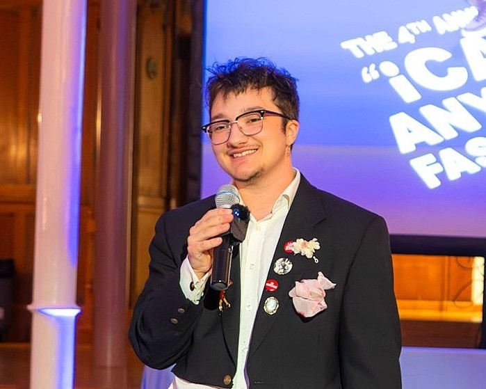 A person stands on stage holding a microphone and smiling, dressed in a dark suit jacket decorated with pins and fabric flowers, in front of a projection reading “The 4th Annual ‘iCAN Do Anything’ Fashion Show.”