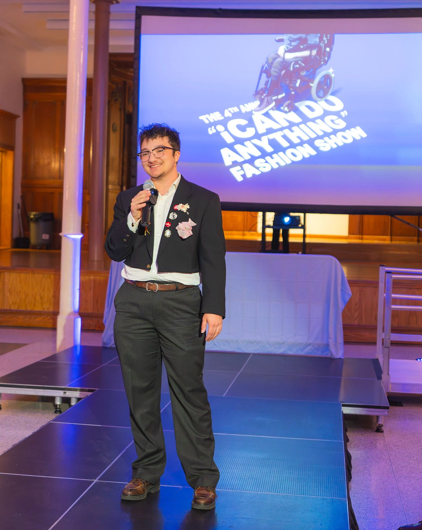 A person stands on a runway holding a microphone and smiling. They wear a dark suit with a white shirt and decorative pins and flowers on the jacket. A large projection screen behind them reads “The 4th Annual ‘I Can Do Anything’ Fashion Show.”