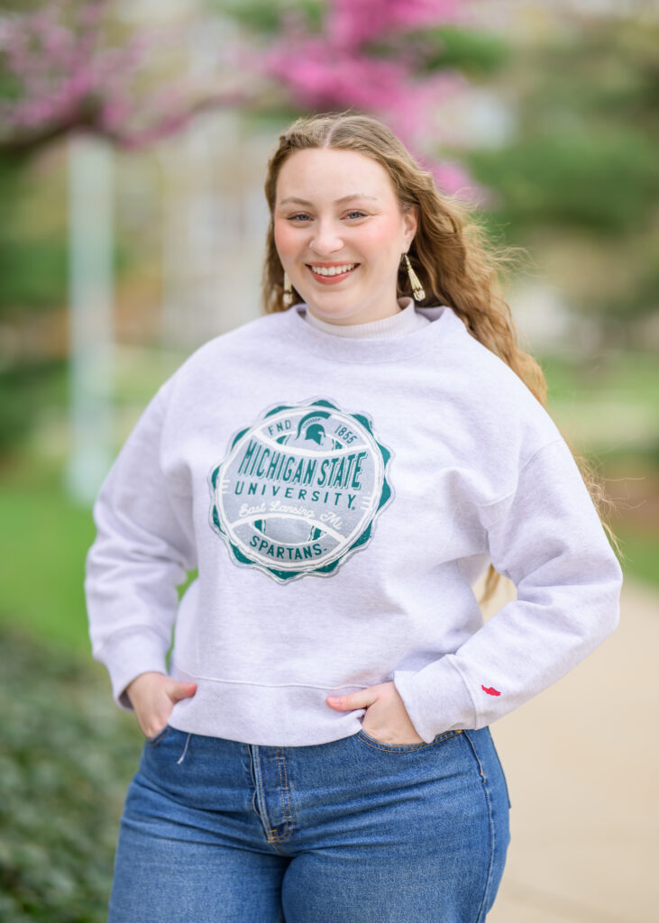A person smiles outdoors while wearing a light gray Michigan State University sweatshirt. They have long blonde hair. Pink flowering trees and green foliage are visible in the background.