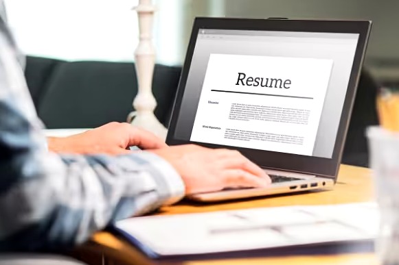 A person types on a laptop displaying a document titled “Resume,” with sections for education and work experience visible on the screen.
