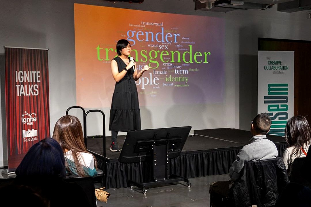 Alex Guo stands on stage speaking into a microphone during Ignite Talks MSU at the MSU Museum CoLab Studio. A colorful word cloud with terms like “transgender,” “gender,” and “identity” is projected behind her as audience members watch from their seats.