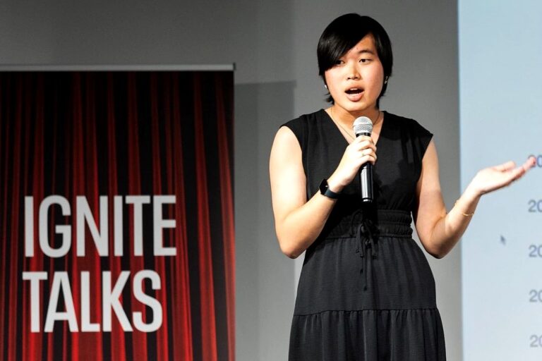 An Asian woman with black short hair and an all-black sleeveless dress with a v-neck, holding a silver microphone and wearing a black watch. She is on a stage with a banner display behind her that says "Ignite Talks" in white letters on a red background.