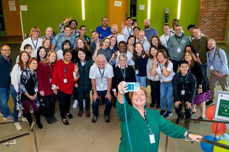 A large, diverse group of faculty, alumni, students, and guests pose together in a bright atrium with green walls during Michigan State University’s Second Language Studies Ph.D. program 20th anniversary celebration. Many wear MSU name badges and SLS-branded shirts.