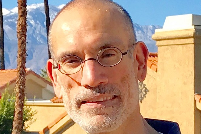 A man with short hair, a graying beard, and round glasses stands outdoors in bright sunlight. Behind him are tan stucco buildings, tall palm trees, and snow-capped mountains.