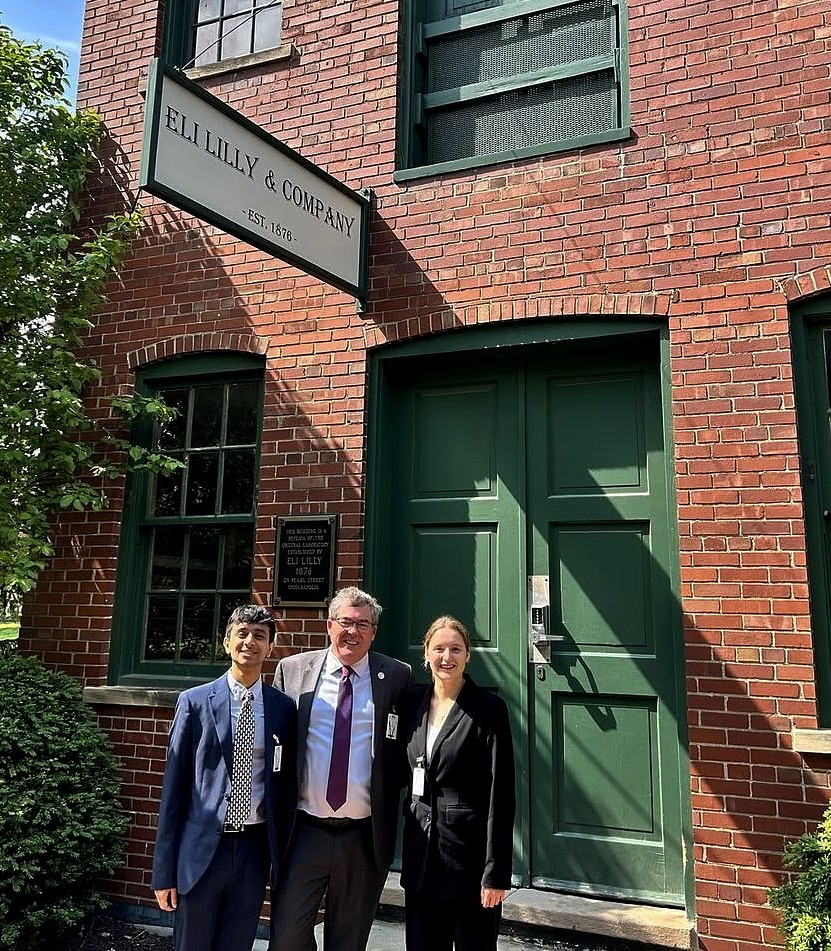 Three people stand in front of a historic red brick building with tall green doors and windows. A hanging sign above them reads “Eli Lilly & Company – Est. 1876.” The person on the left wears a blue suit and patterned tie, the person in the middle wears a suit with a burgundy tie, and the person on the right wears a black suit. All three are smiling.