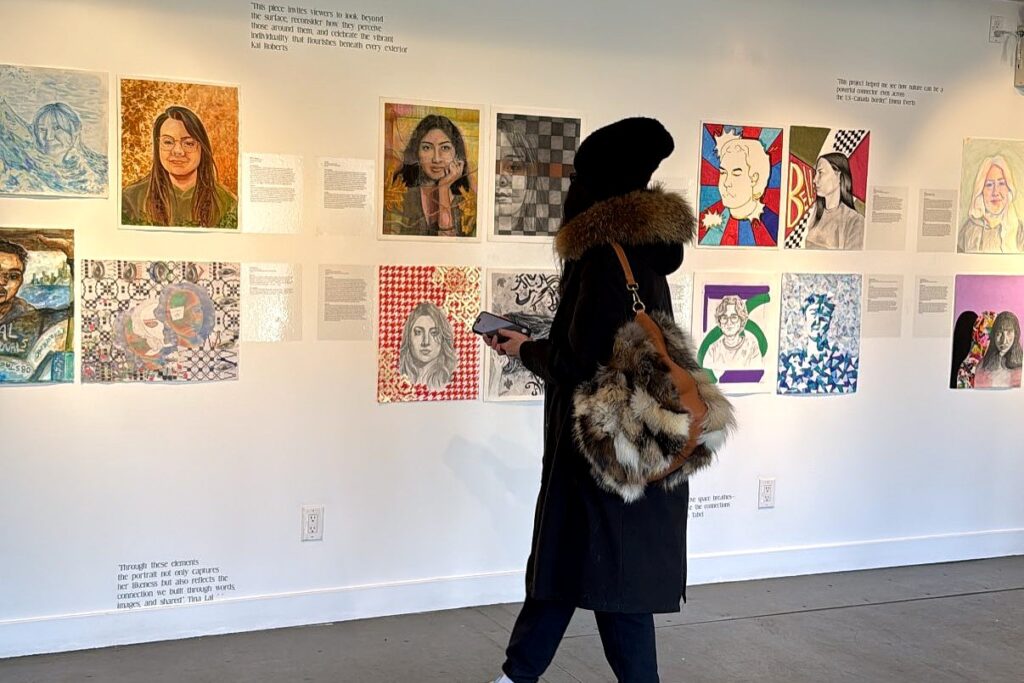 Woman in a long black winter coat and black knit hat carrying a fake fur covered purse and a cell phone looking at a wall of painted and drawn portraits at a gallery.