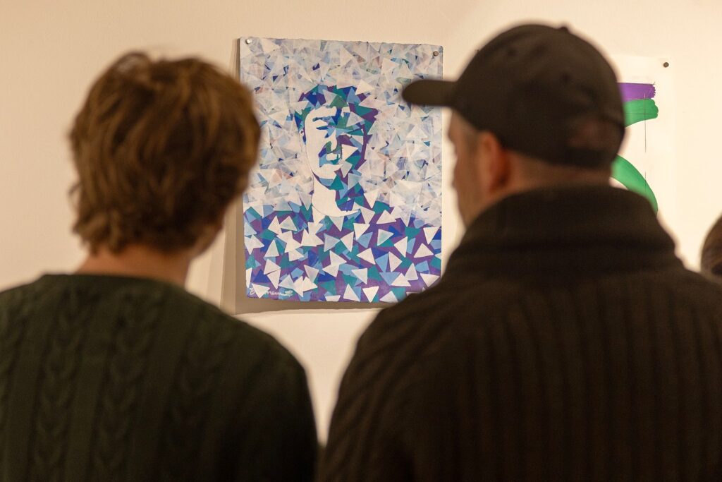 Two people seen from behind look at a portrait made of layered blue, purple, and white triangular shapes forming the outline of a face.
