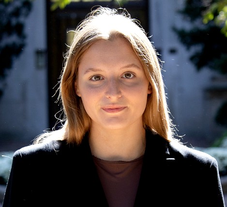 A person stands outdoors in direct sunlight, facing the camera with a neutral expression. They have long, light-colored hair and wear a dark blazer over a high-neck top. The background is softly blurred with trees and building details visible.