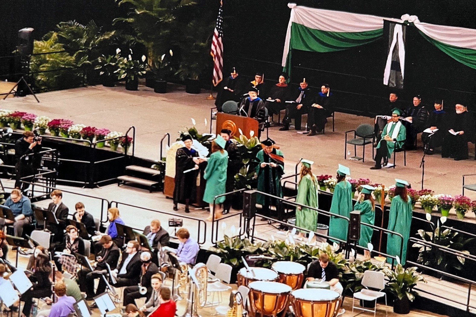 Commencement ceremony with graduates in green caps and gowns walking across a stage to receive diplomas. Faculty members sit on stage under green and white drapery, and a band performs in the foreground.