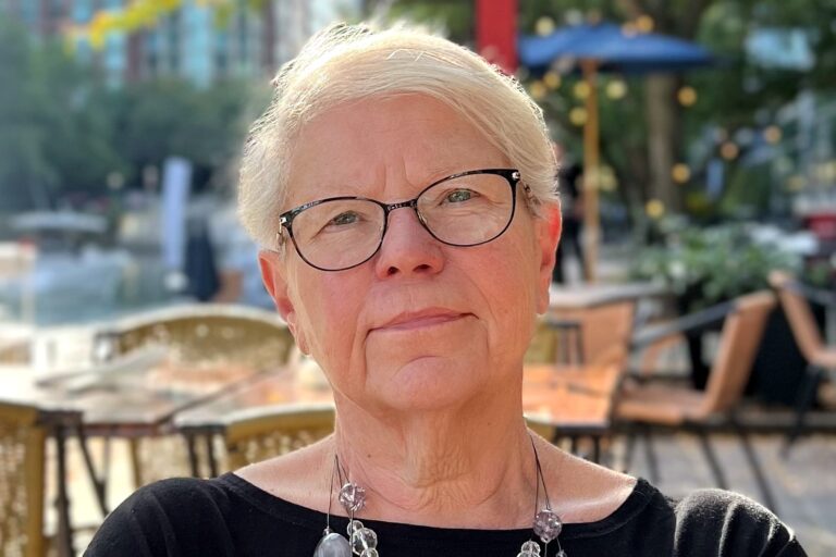 Close-up outdoor portrait of a woman with short light hair and glasses, wearing a black top and a layered beaded necklace. Blurred café tables and trees appear in the background.
