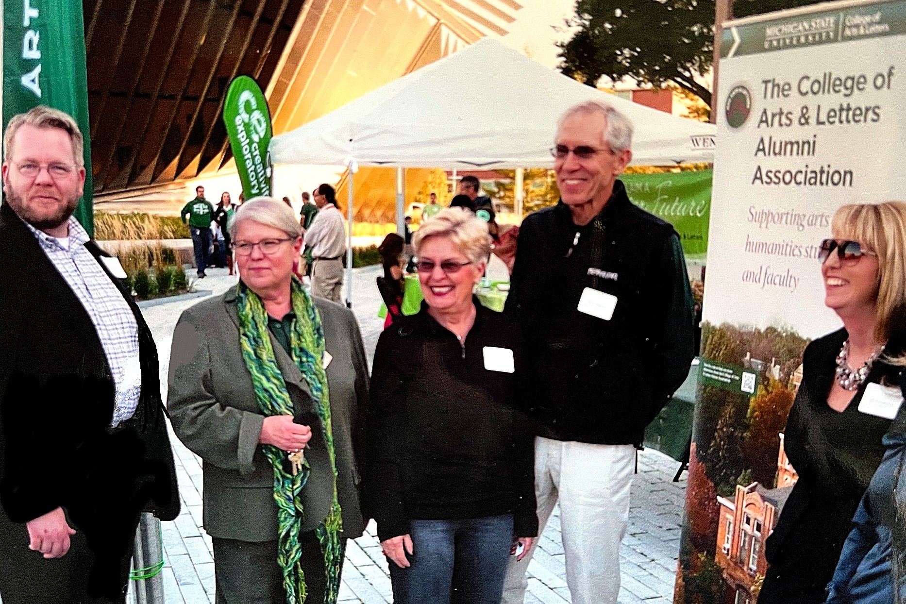 A group of five adults standing outdoors at an MSU event, with campus tents and green MSU-branded banners in the background. All are wearing nametags and talking together near a College of Arts & Letters Alumni Association display sign.