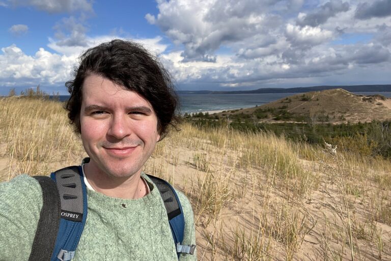 A person with black hair stands smiling on a windswept sand dune overlooking Lake Michigan, wearing a green sweater and backpack. Behind them, rolling dunes, grasses, and a blue sky with scattered clouds.