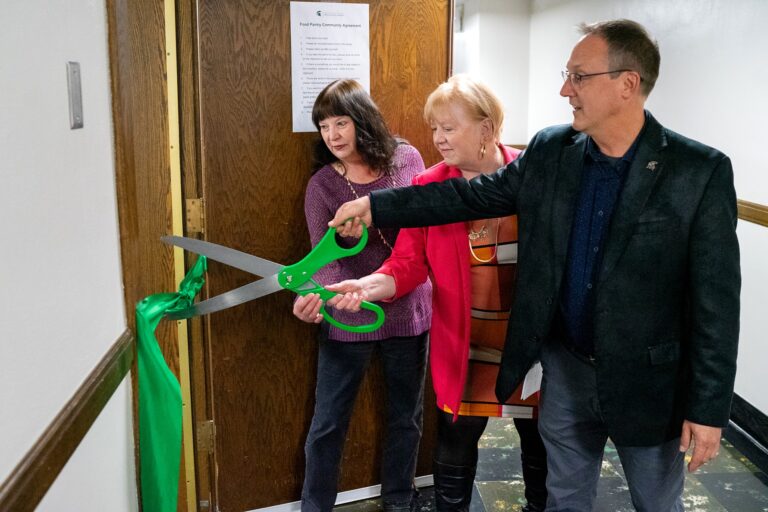 Three people stand in a hallway beside a door with a green ribbon tied across it. Two individuals hold large green ceremonial scissors together while a third person stands beside them, watching the cut.