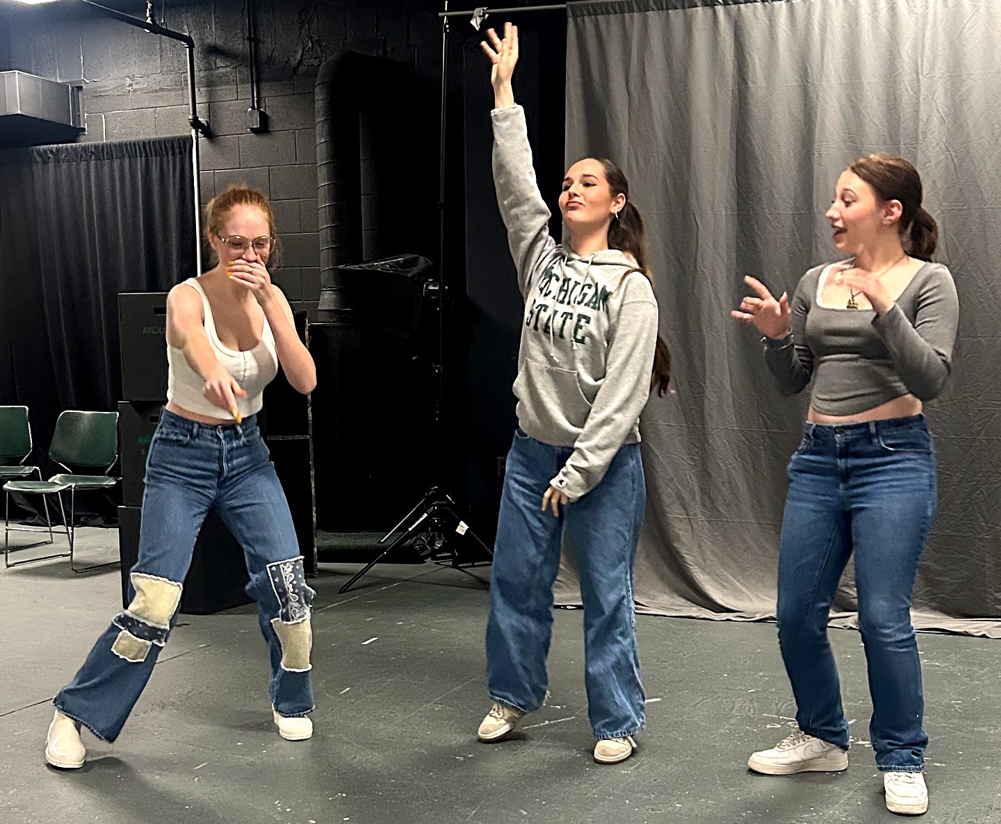 Three students rehearse an improv exercise in a studio. One student in a white tank top and patched jeans leans forward, another in an MSU sweatshirt raises an arm, and a third reacts. Lighting equipment and chairs are visible in the background