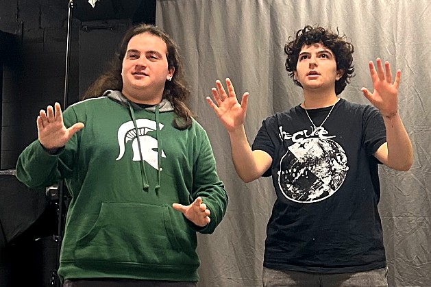 Two students stand side by side in an improv rehearsal, each holding their hands up mid-gesture as if responding to an imagined situation. One wears an MSU Spartan hoodie and the other a black graphic T-shirt.