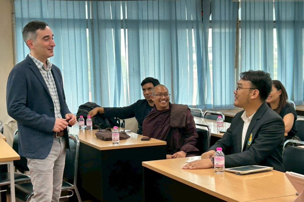 A teacher talks with four adult students who are sitting at two rows of tables.